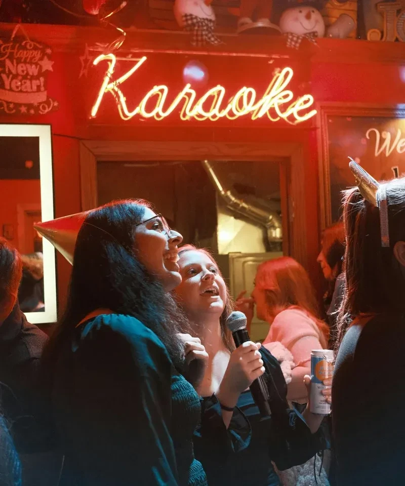Young women sing karaoke under a red neon "Karaoke" sign.