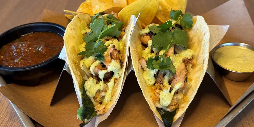Two filled soft tacos with green sauce, cilantro, salsa, and chips.