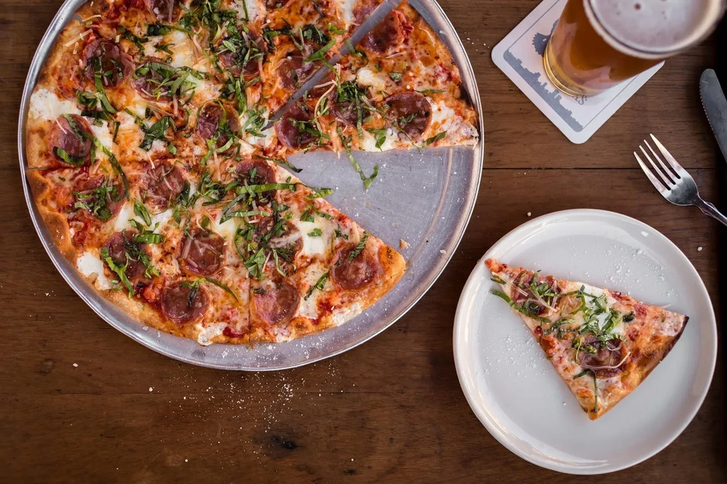 Overhead: pepperoni pizza slice, beer, fork, knife on wooden table.