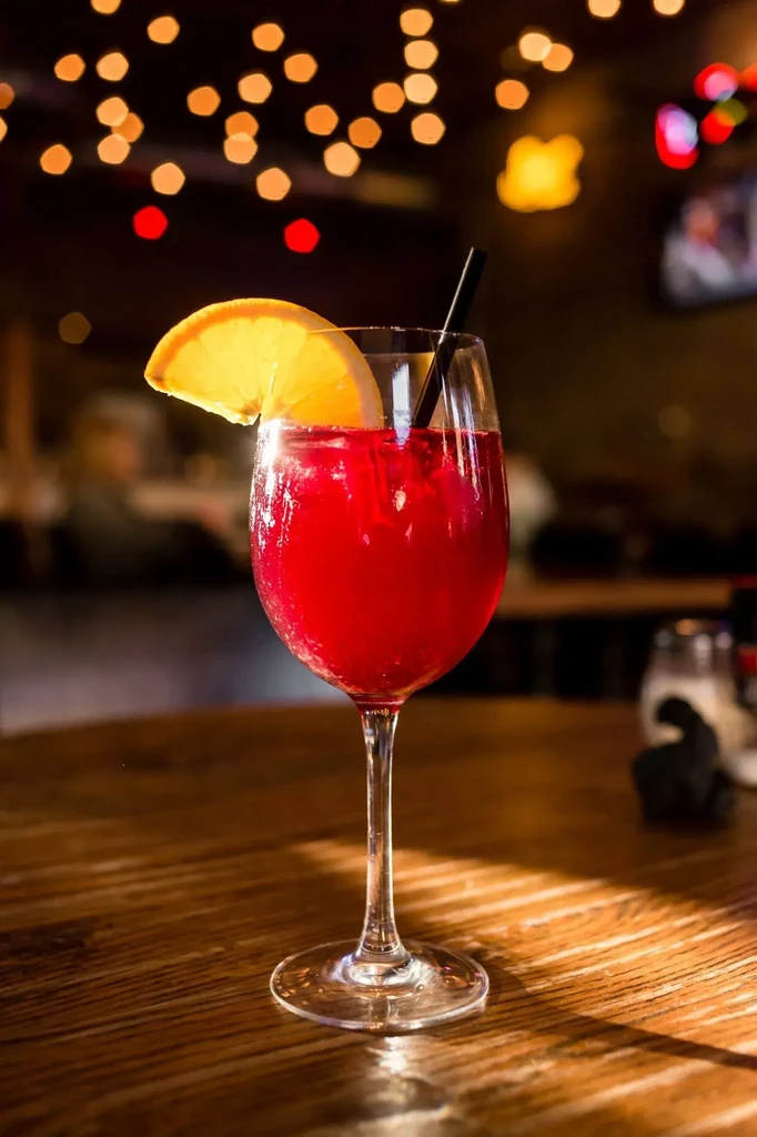 Red drink in wine glass with orange, straw, ice on wood.