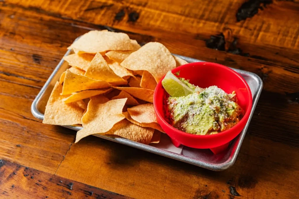 Tortilla chips and guacamole on a silver tray with lime, wooden table.