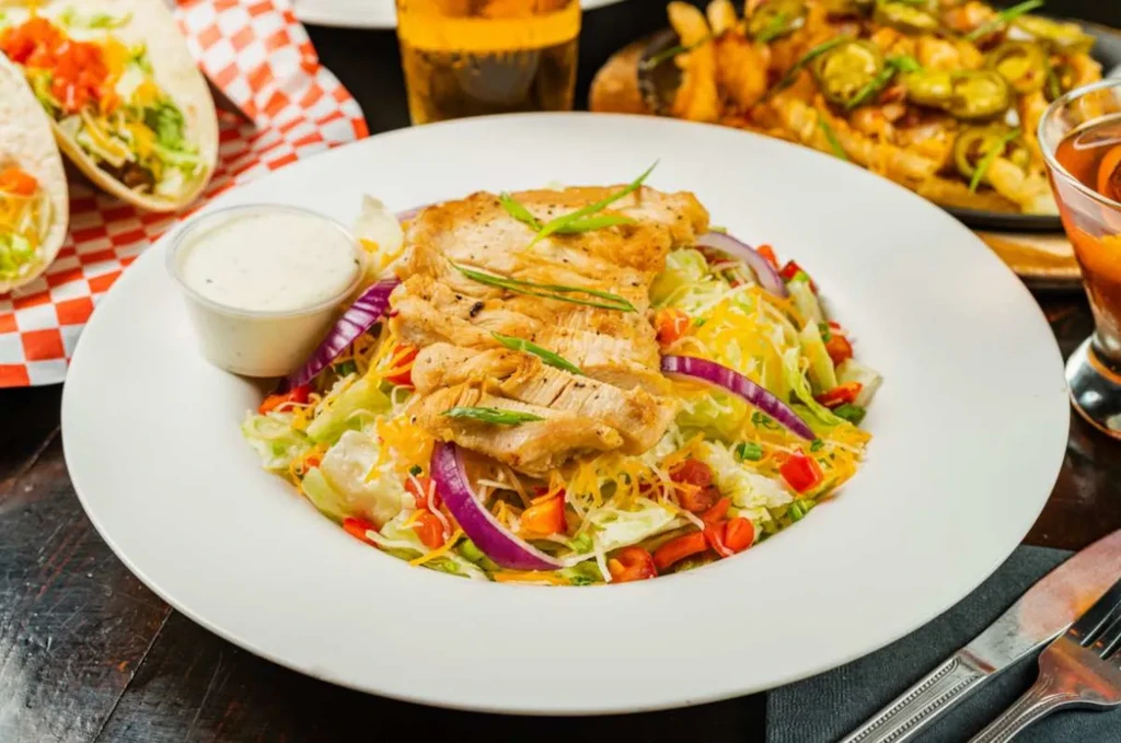 Plated grilled chicken salad with ranch, tacos, beer, and nachos.