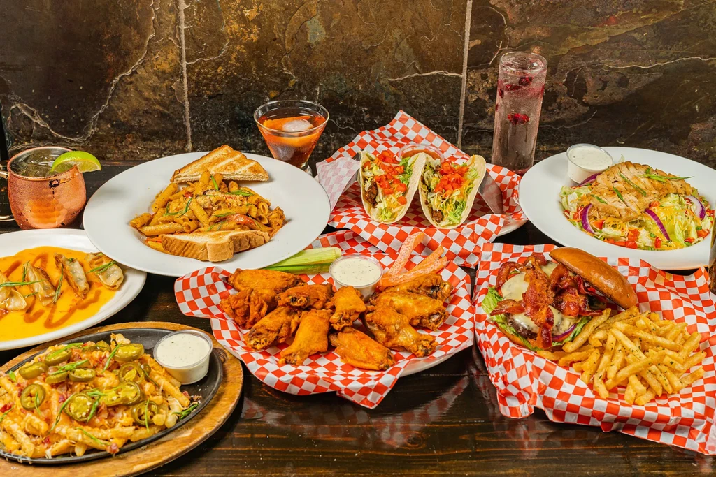 Spread of pasta, tacos, burger, wings, fries, and drinks.