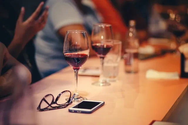 Red wine, eyeglasses, phone on peach table in dimly lit restaurant.