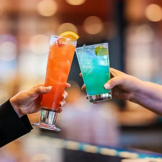 Two hands clinking tall orange and short blue cocktails with garnishes.