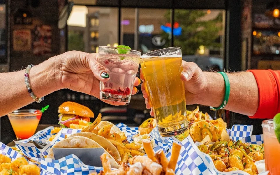 Two hands toast a cocktail and beer over a spread of bar food.
