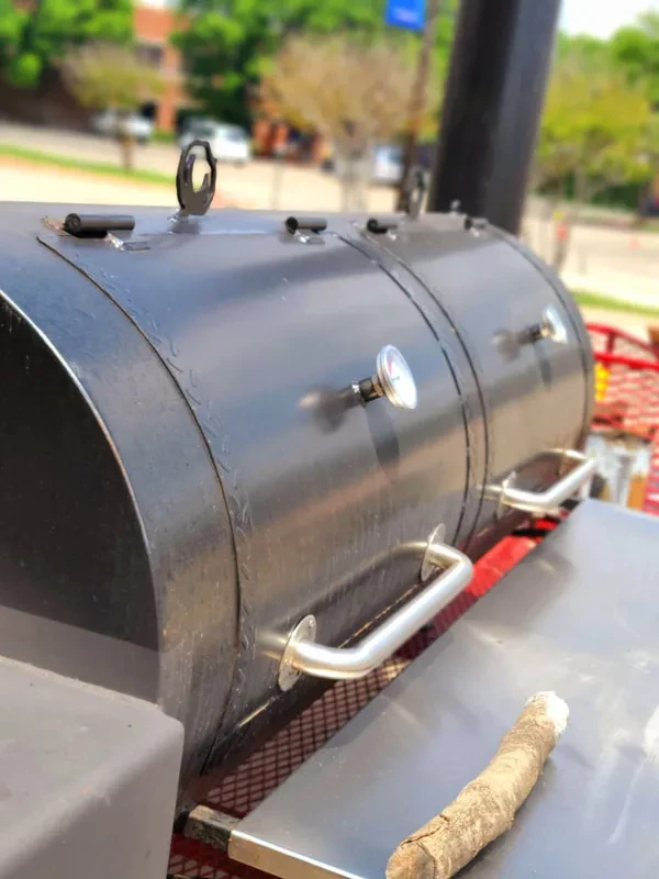 Large black barrel smoker with wood chunk on metal workspace.