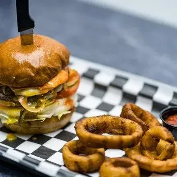 Large burger with knife, crispy onion rings, and red dipping sauce.