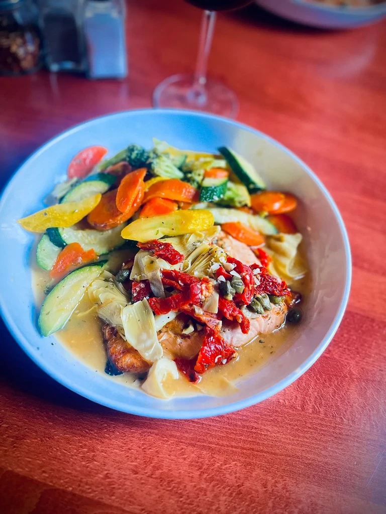Cooked fish with sun-dried tomatoes, artichokes, capers, vegetables in a bowl.