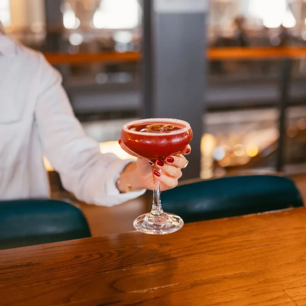 Hand with red nails holding a red cocktail on a wooden bar.