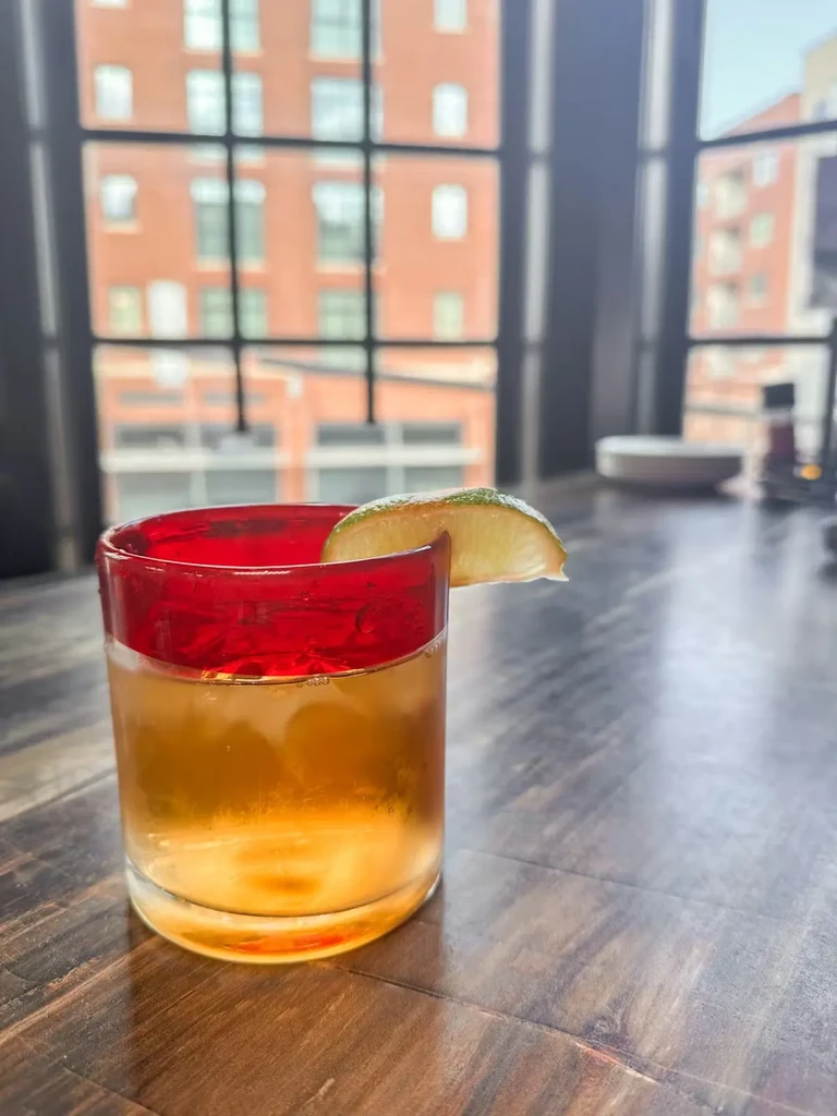 Amber drink with ice, red rim, and lime, by an urban window.