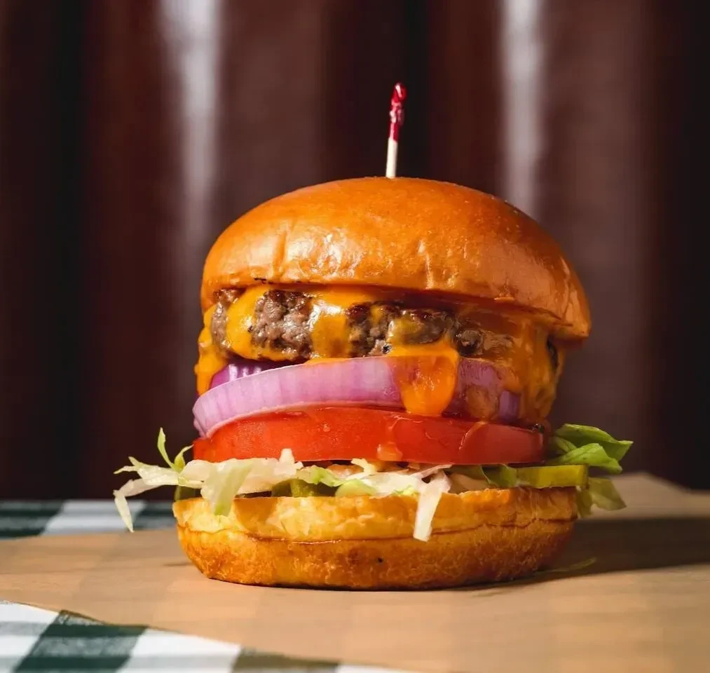 Well-lit close-up of a tall gourmet burger with melted cheese and vegetables.