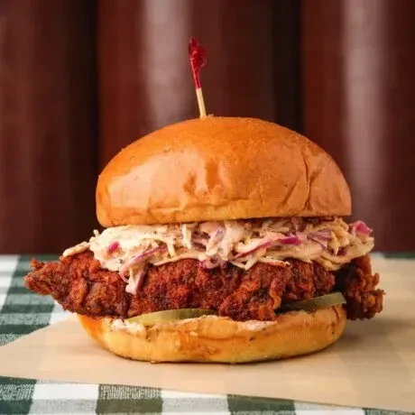 Crispy fried chicken sandwich with coleslaw, pickles, on checkered cloth.