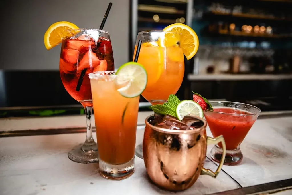 Five vibrantly colored cocktails with garnishes on a bar.