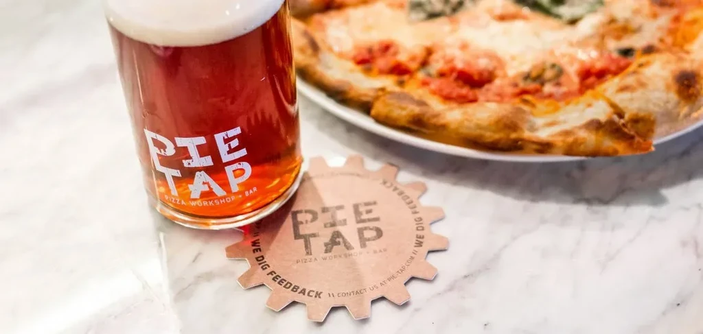 Amber Pie Tap beer, branded coaster, and blurred pizza.