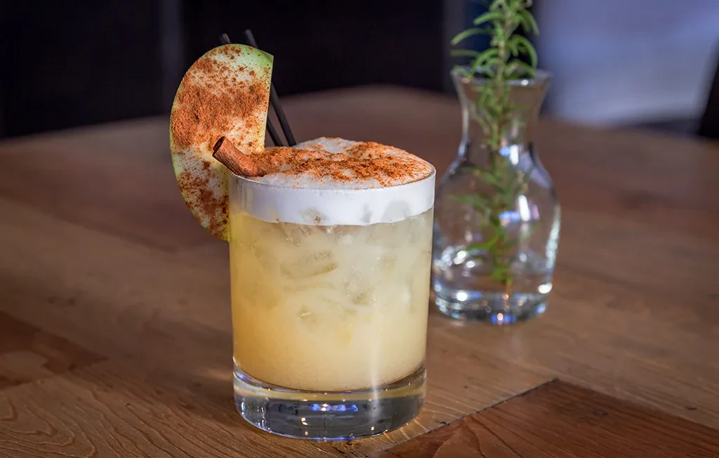 Amber cocktail with spiced foam, green apple slice, and cinnamon stick.