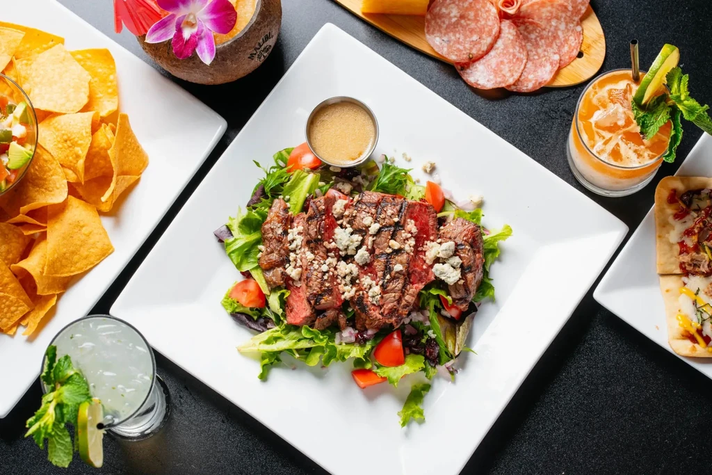 Top-down view: steak salad, chips, meats, cocktails, and tropical drinks.