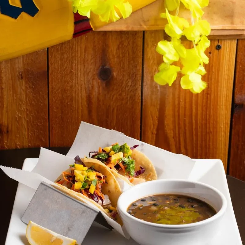 Two vibrant tacos, black bean soup, lemon, and yellow flower garlands.