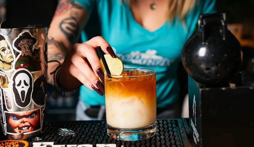 Bartender's tattooed hand garnishes a layered drink at a horror-themed bar.