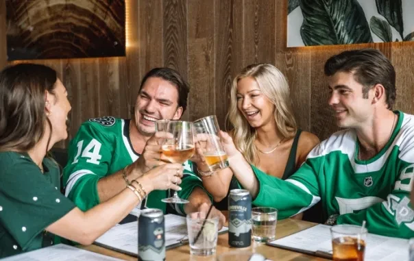 Four young adults, two in hockey jerseys, toast drinks in a restaurant.