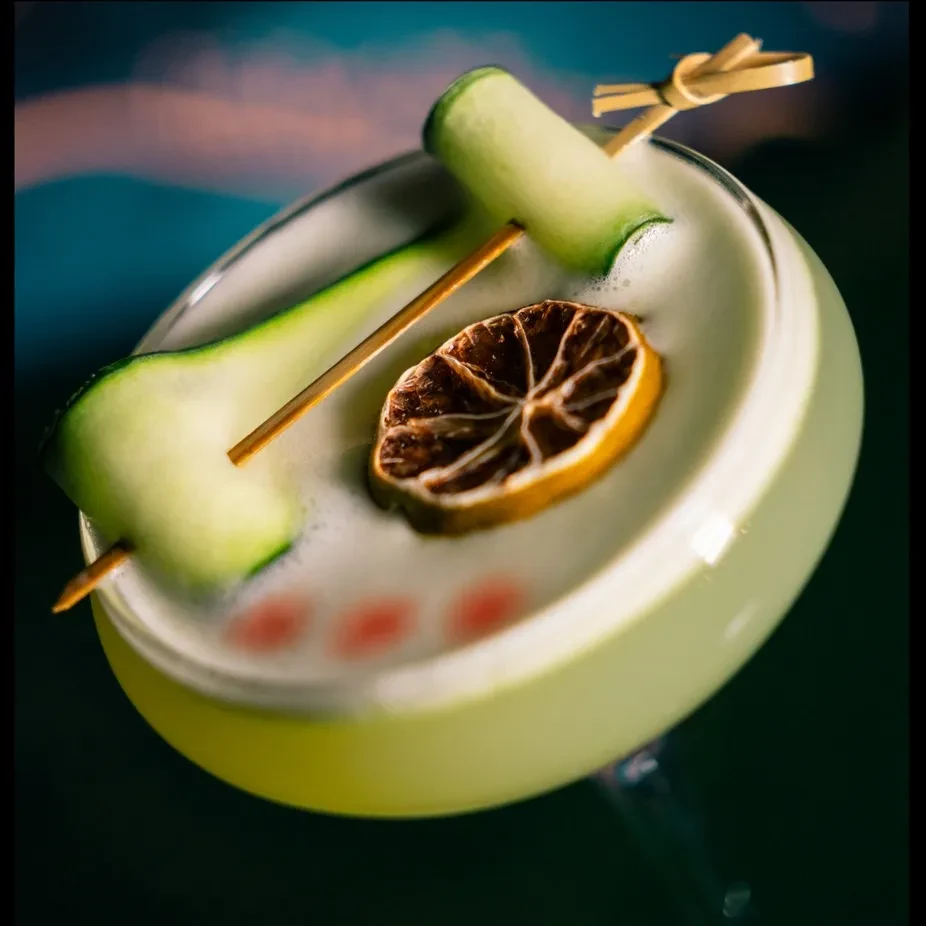 Martini cocktail with cucumber skewer, dried citrus, and red dots on foam.