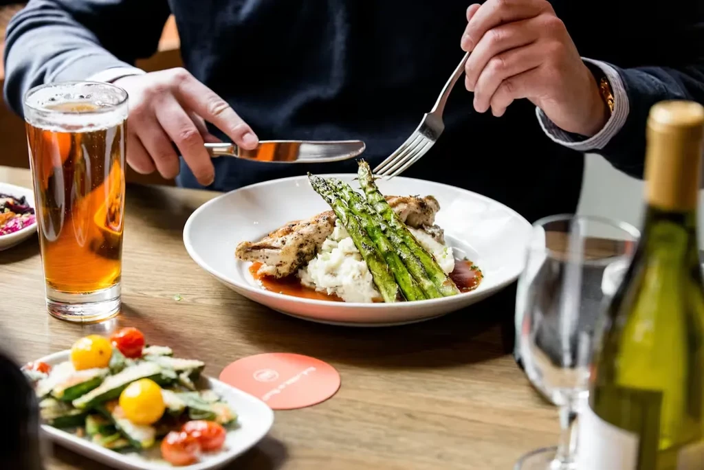 Person cutting grilled chicken, mashed potatoes, asparagus, and beer on wooden table.