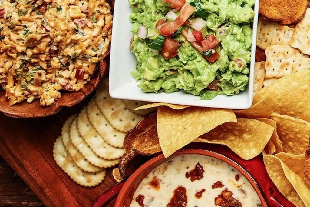 Top-down view of three dips, crackers, and chips on a rustic board.