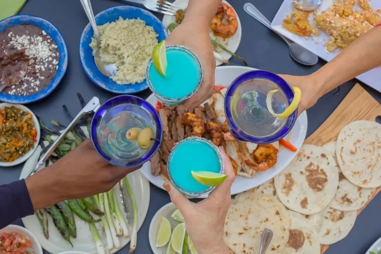 Overhead: Multiple hands toast colorful drinks and diverse food.