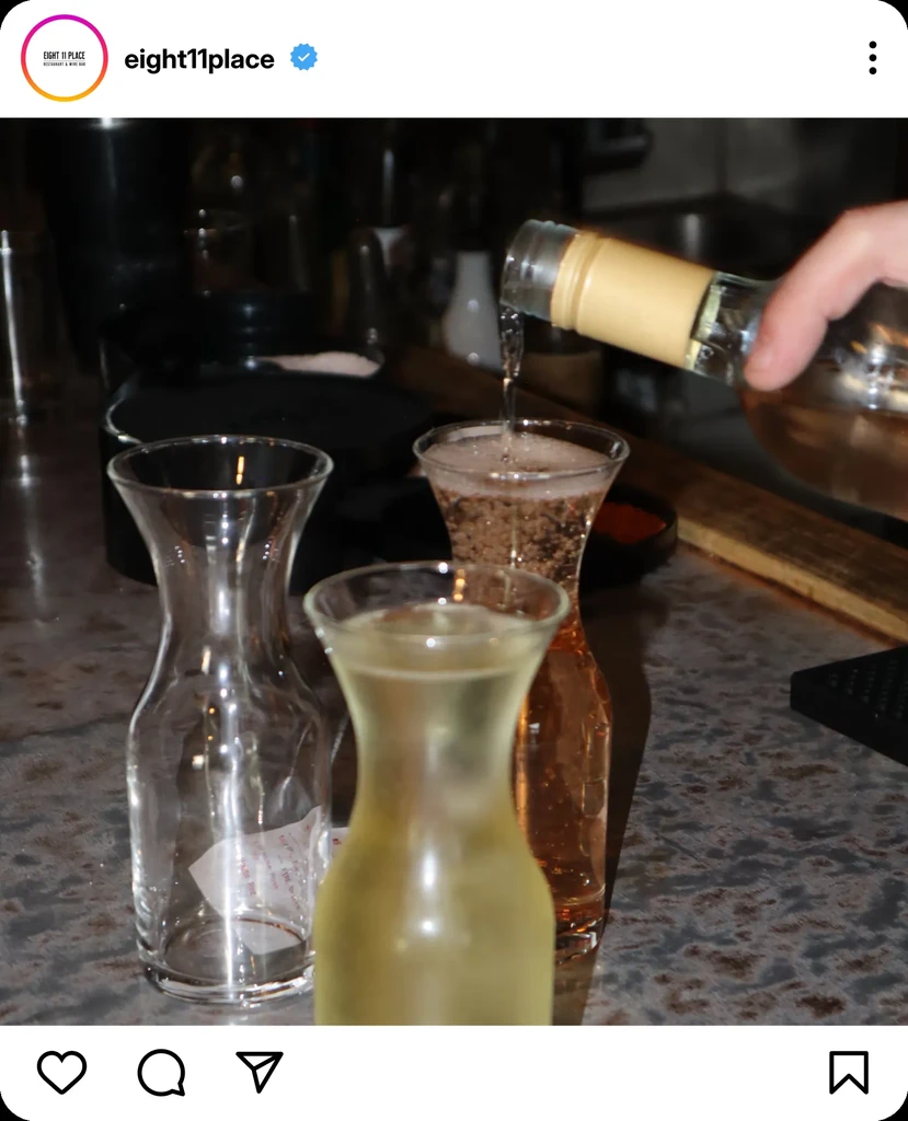 Hand pours effervescent beverage into carafe among others on bar counter.