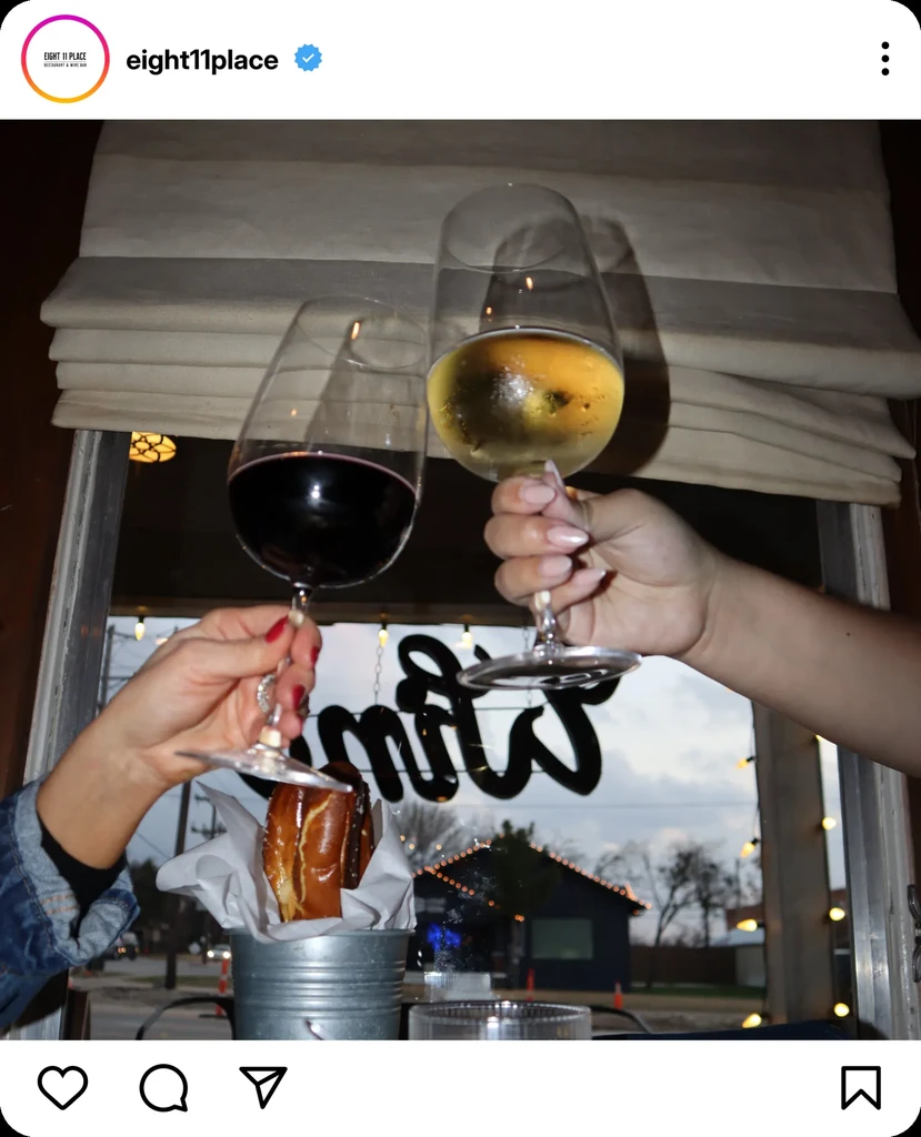 Two hands toast red and white wine, a pretzel below, at dusk.