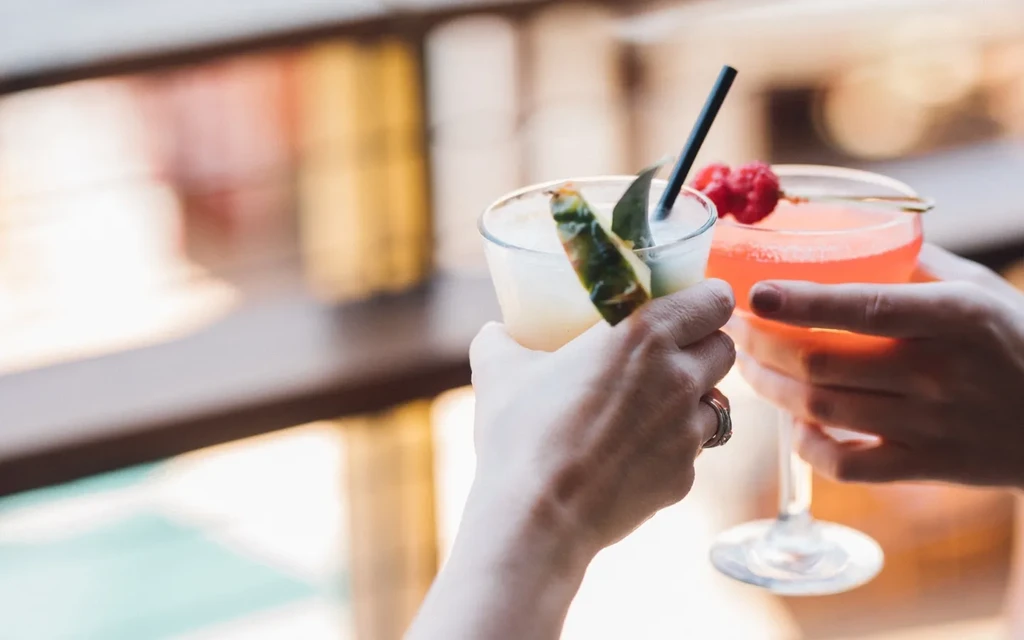 Hands toasting two cocktails: creamy pineapple drink and pink raspberry coupe.
