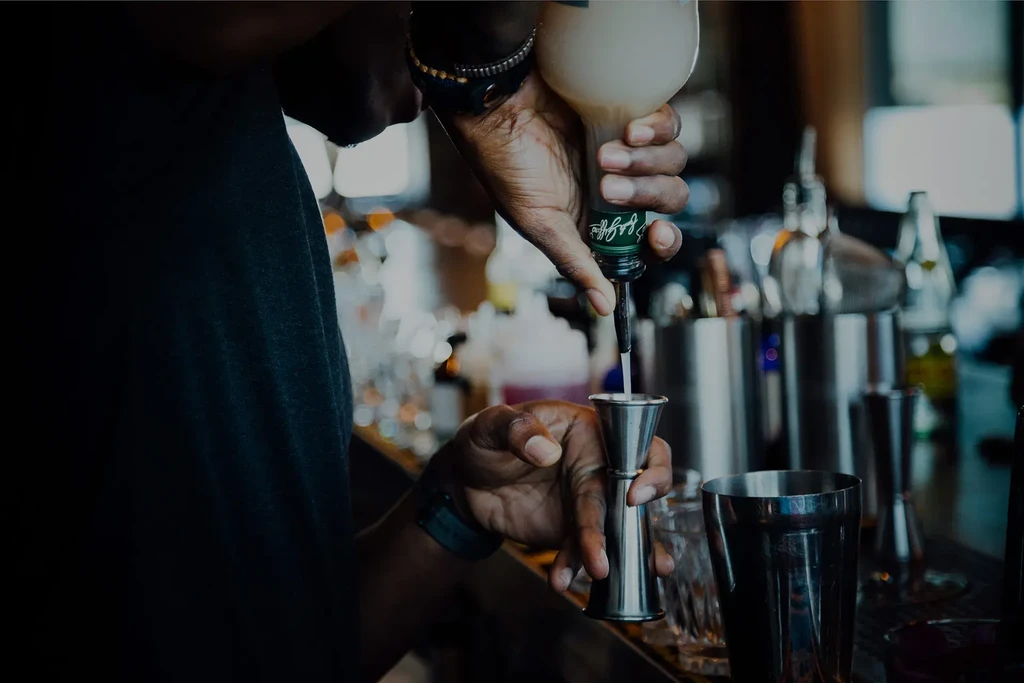 Hands pour light liquid from squeeze bottle into metal jigger.