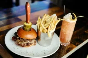 Gourmet burger, fries, and pink cocktail on a patterned wooden table.