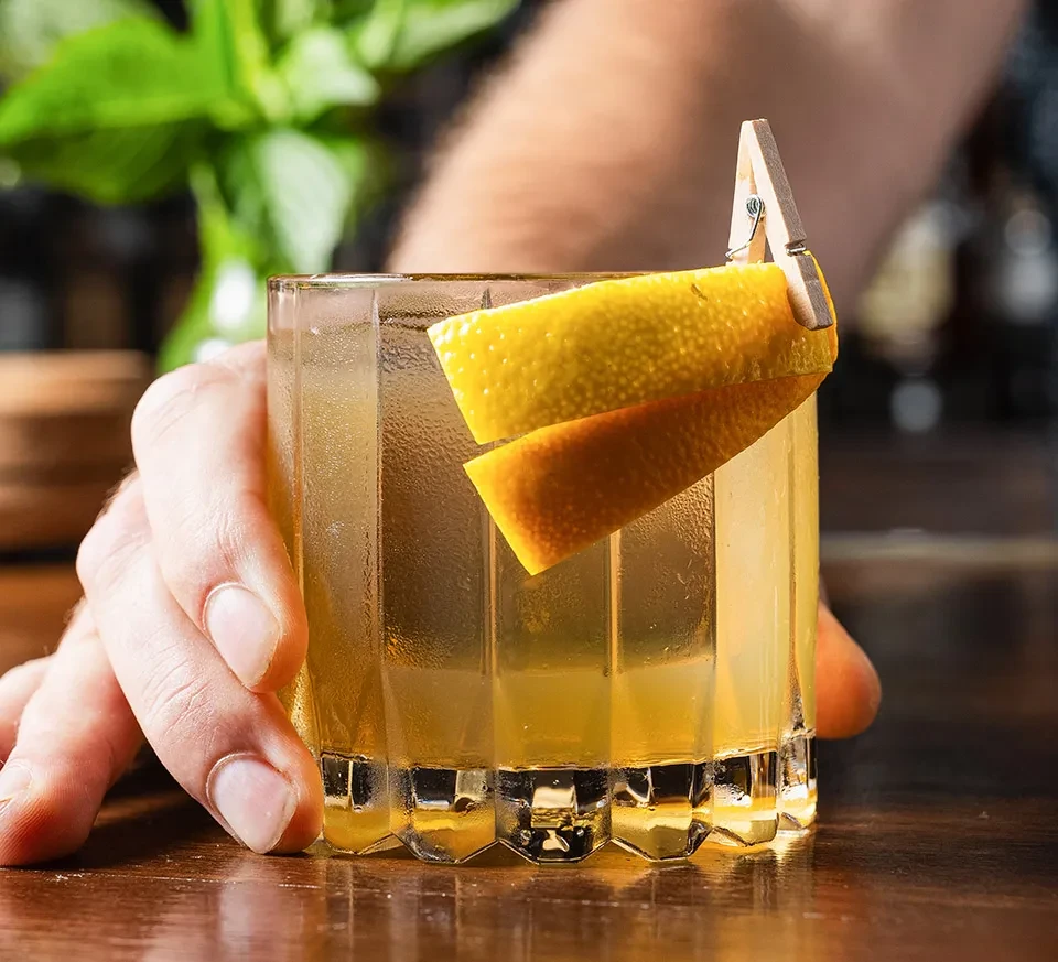 Hand holding a faceted glass with a golden drink and citrus garnish.
