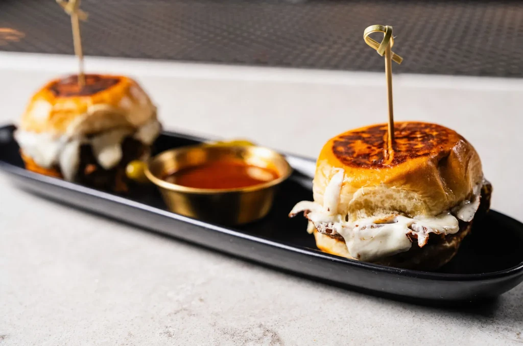 Two mini burgers with melted cheese and dipping sauce on a black tray.