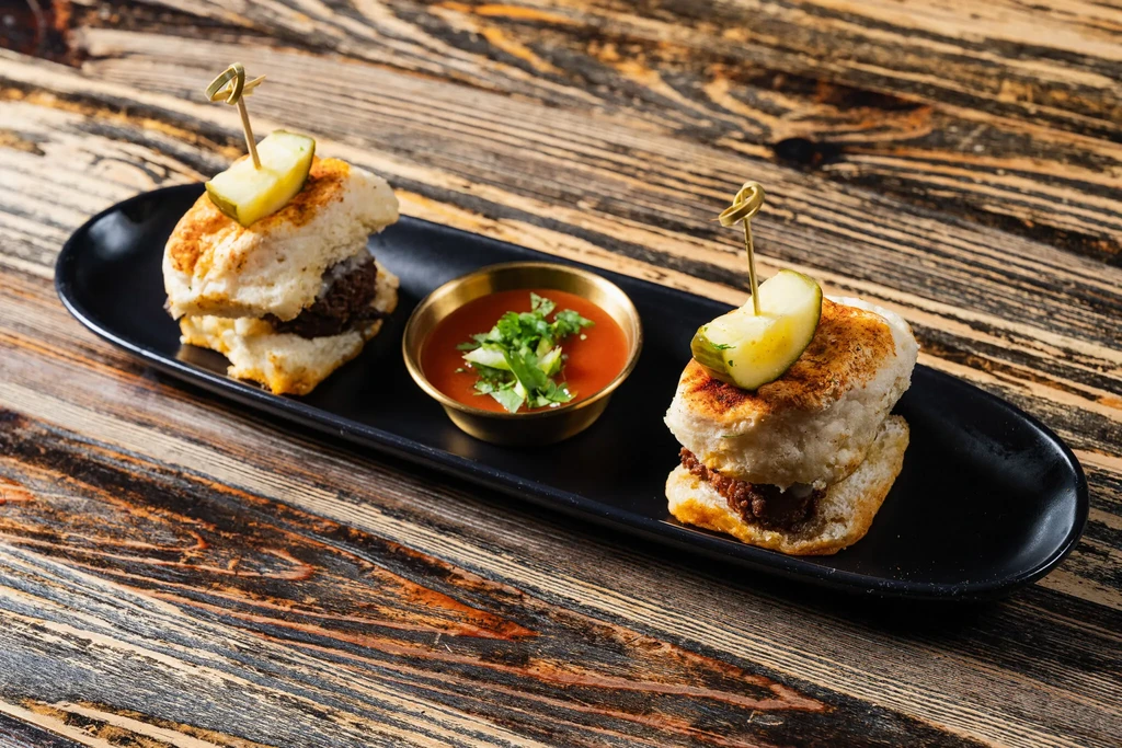 Two sliders with pickles, herb sauce, and skewers on a black plate.