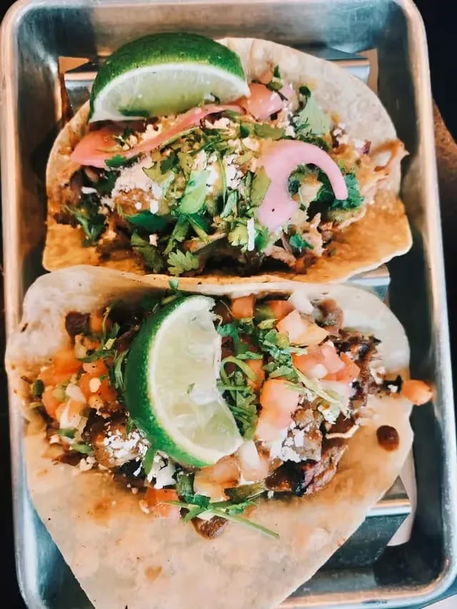 Two gourmet tacos with meat, cheese, and garnishes in a metal tray.