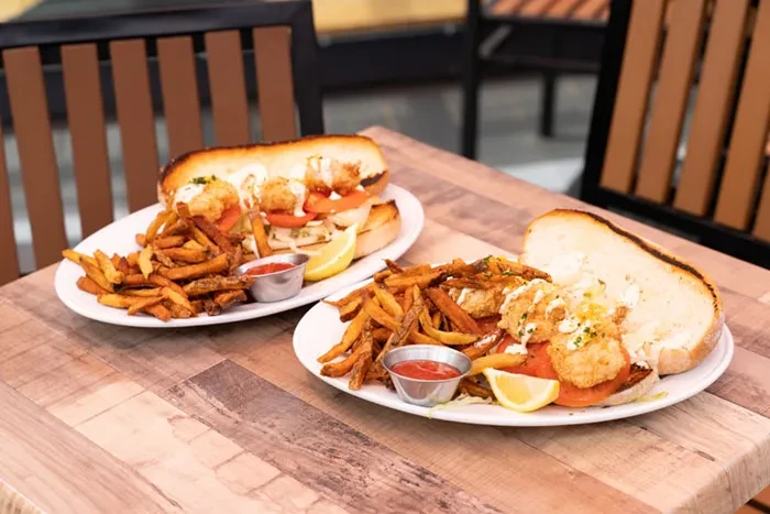 Two plates of fried shrimp po'boys with fries on a wooden table.