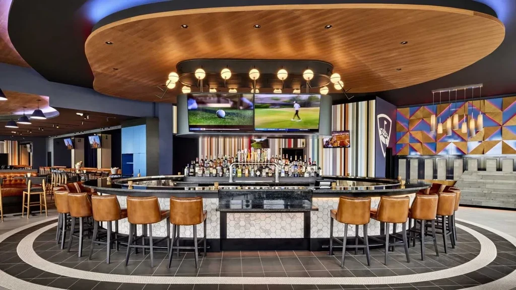 Modern sports bar with circular bar, screens showing golf, patterned floor.