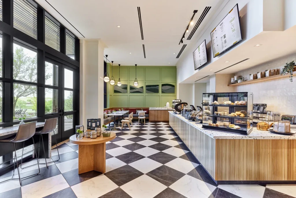 Bright modern cafe with bakery display, checkered floor, and green views.
