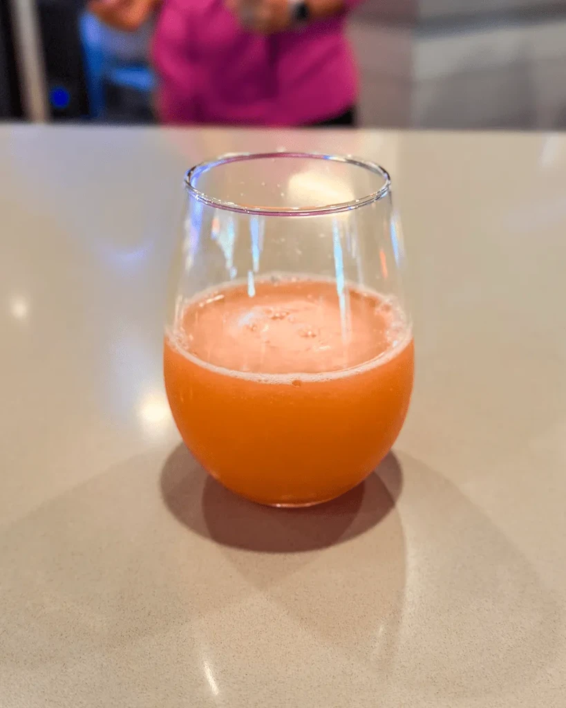 Frothy orange drink in a stemless glass; blurred person behind.