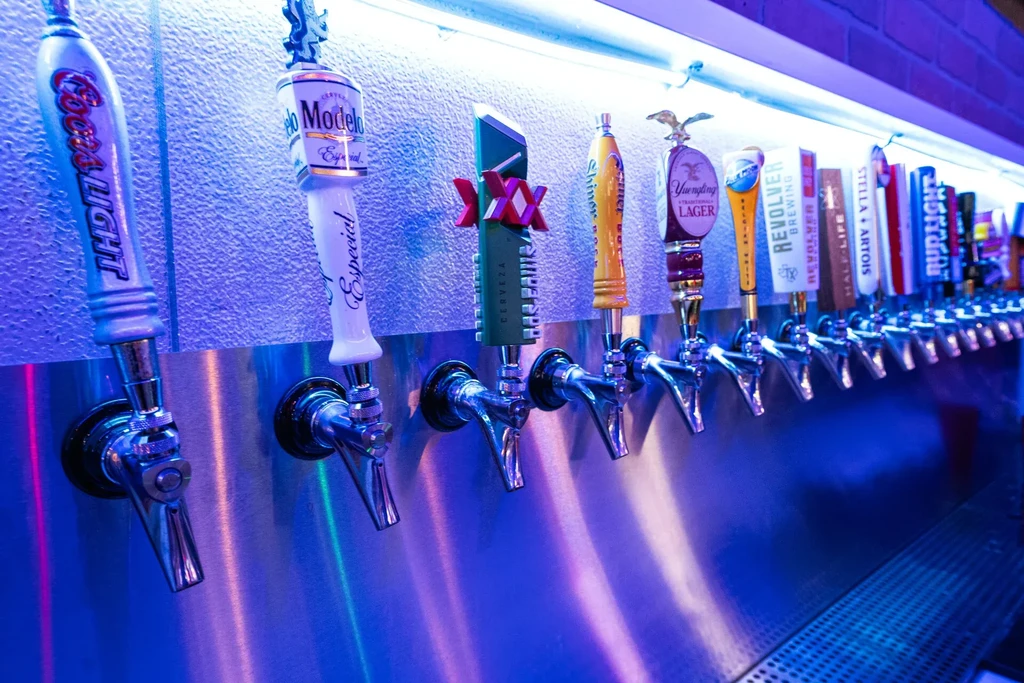 Shiny beer taps with brand logos glow blue in a bar.