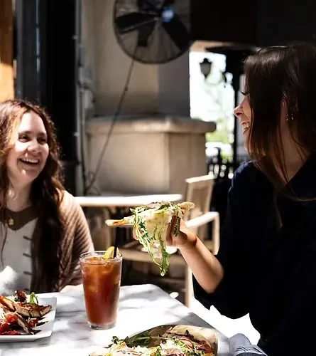 Two smiling women dining outdoors; one holds up an arugula pizza slice.