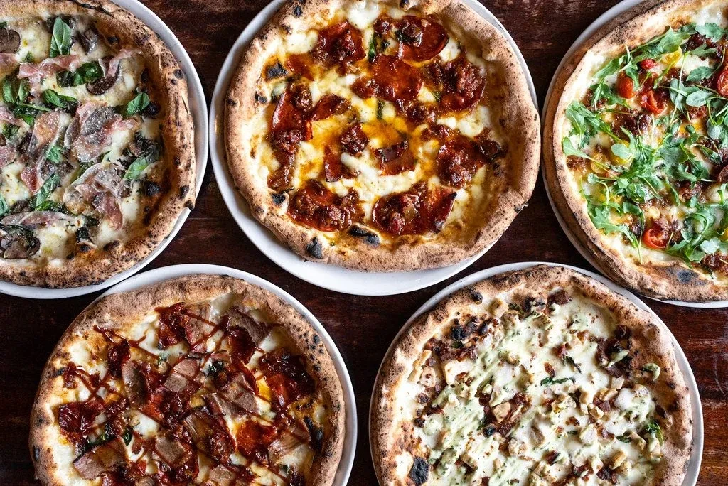 Five distinct artisan pizzas with varied toppings on a dark wooden table.