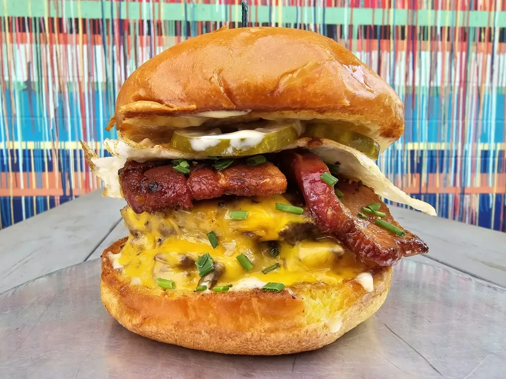 Gourmet burger with two patties, melted cheese, bacon, egg, and pickles.