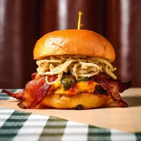 Close-up of a stacked burger with bacon, melted cheese, jalapeños, fried onions.