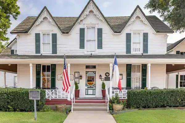 White Victorian building, green shutters, porch, American and Texas flags, historical marker.