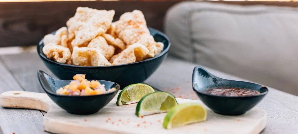 Crispy chicharrón, two dips, lime, and spices on a wooden board.
