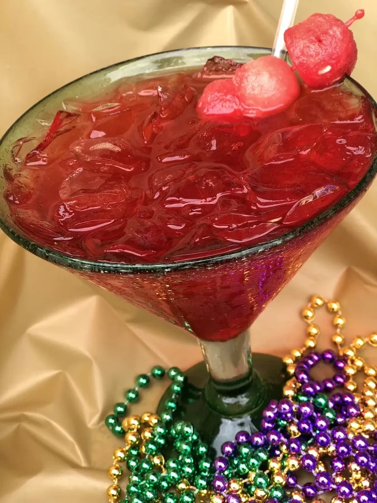 Red iced cocktail in green-stemmed glass, fruit, Mardi Gras beads.