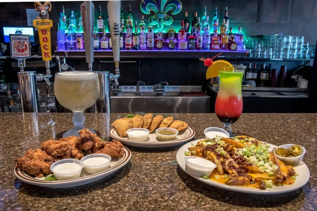 Fried chicken, empanadas, loaded fries, beer, and cocktail at a lit bar.